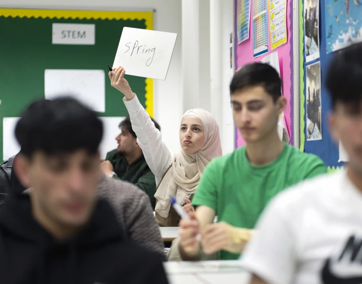 A female student in a beige hijab holds up a small whiteboard with the word "Spring" written on it, participating in a classroom activity. Other students are in the foreground, slightly out of focus. A female student in a beige hijab holds up a small whiteboard with the word "Spring" written on it, participating in a classroom activity. Other students are in the foreground, slightly out of focus.